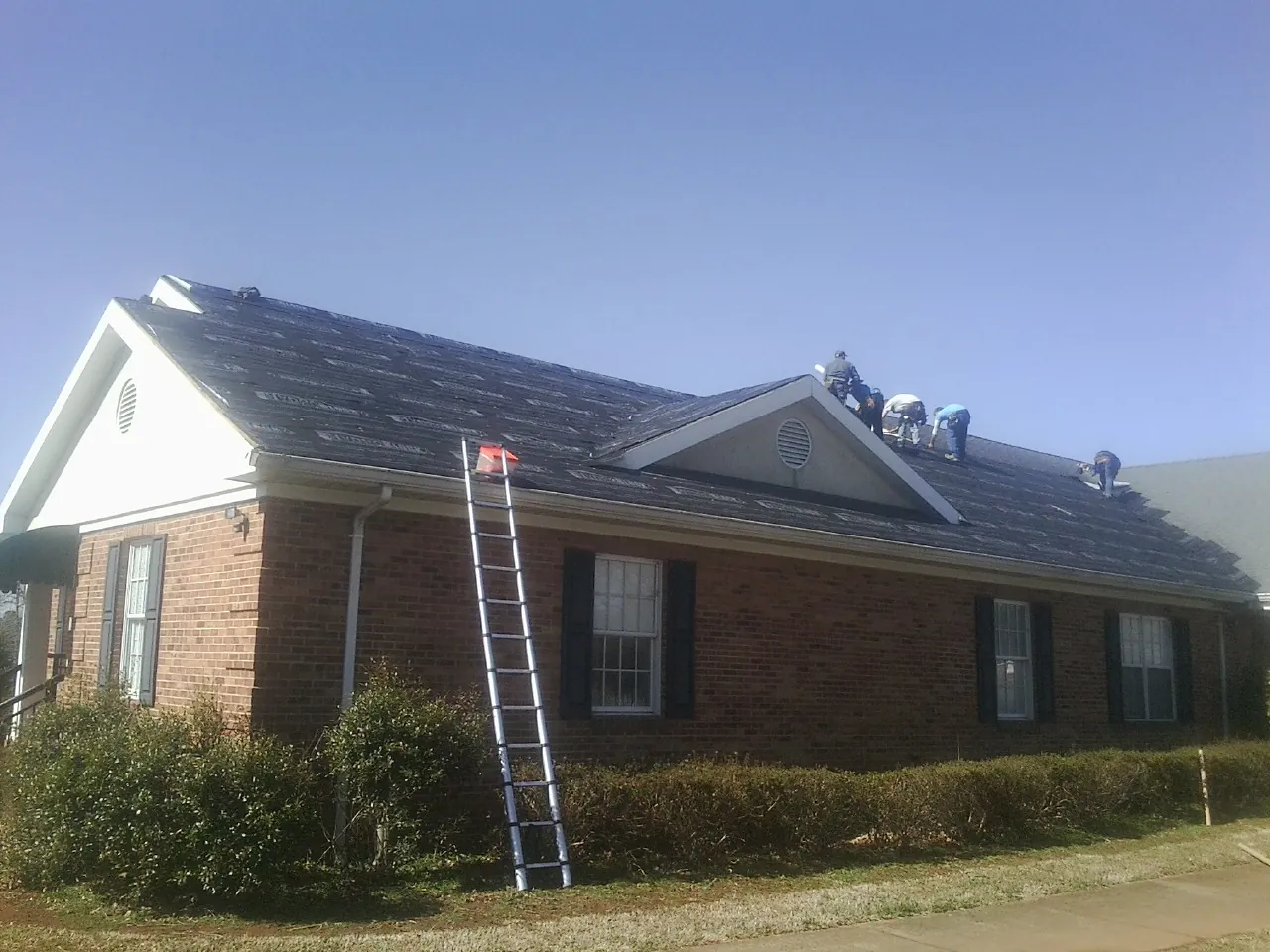 Crew tearing shingles off a brick ranch roof with a ladder set — Metal Roof Repair work by our crew in Walton